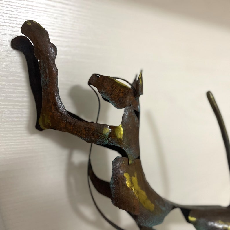 Op de afbeelding: Een roestig metalen sculptuur van een kat met gouden en blauwe verfaccenten. De kat staat in een speelse pose met zijn voorpoot omhoog.