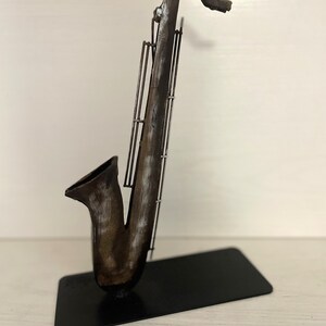 May include: A metal sculpture of a saxophone with a distressed brown and white paint finish. The saxophone is standing on a black metal base.