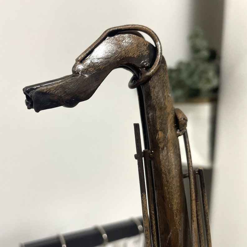 May include: A metal sculpture of a dog's head with a long, thin neck. The sculpture is painted in a brown and black color scheme, giving it a rustic look.