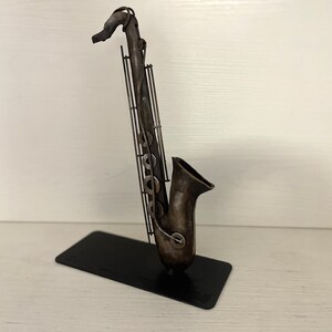 May include: A metal sculpture of a saxophone in a dark brown finish. The saxophone is standing on a black metal base.