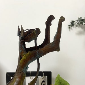 Op de afbeelding: Een metalen sculptuur van een hond met een verweerde, roestige afwerking. De hond staat in een speelse houding met zijn voorpoten omhoog. Het beeld heeft een groene en bruine patina.