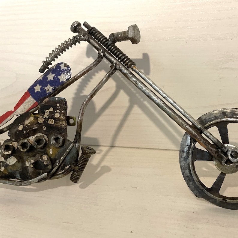 Op de afbeelding: Een metalen sculptuur van een motorfiets met een Amerikaans vlagontwerp op de stoel. De motorfiets is gemaakt van gerecyclede metalen onderdelen en heeft een rustieke, industri&euml;le uitstraling.