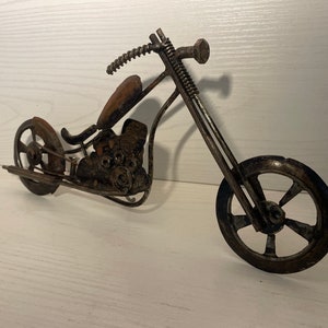 May include: A metal sculpture of a motorcycle, made from recycled materials. The motorcycle is a chopper style with a long front fork and a single seat. The sculpture is painted in a rusty brown color.