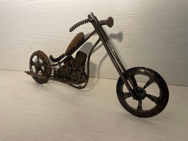 May include: A metal sculpture of a motorcycle, made from recycled materials. The motorcycle is a chopper style with a long front fork and a single seat. The sculpture is painted in a rusty brown color.