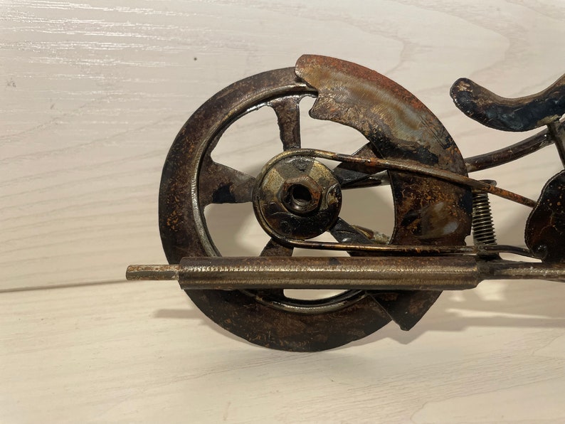 May include: A rusty metal motorcycle sculpture with a single wheel, a seat, and a handlebar. The sculpture is made of recycled metal and has a rustic, industrial look.