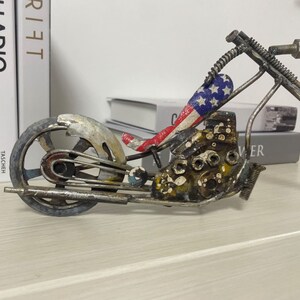 Op de afbeelding: Een metalen motorfiets sculptuur met een rood, wit en blauw Amerikaans vlag ontwerp op de stoel. De motorfiets is gemaakt van gerecycled metaal en heeft een verweerde, roestige afwerking.