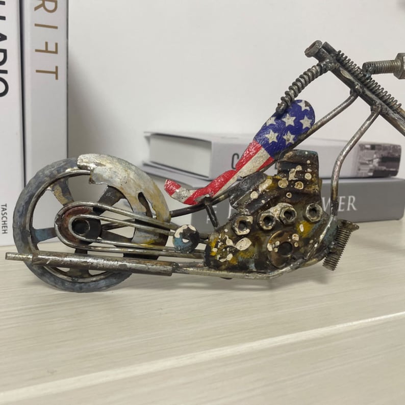 Op de afbeelding: Een metalen motorfiets sculptuur met een rood, wit en blauw Amerikaans vlag ontwerp op de stoel. De motorfiets is gemaakt van gerecycled metaal en heeft een verweerde, roestige afwerking.