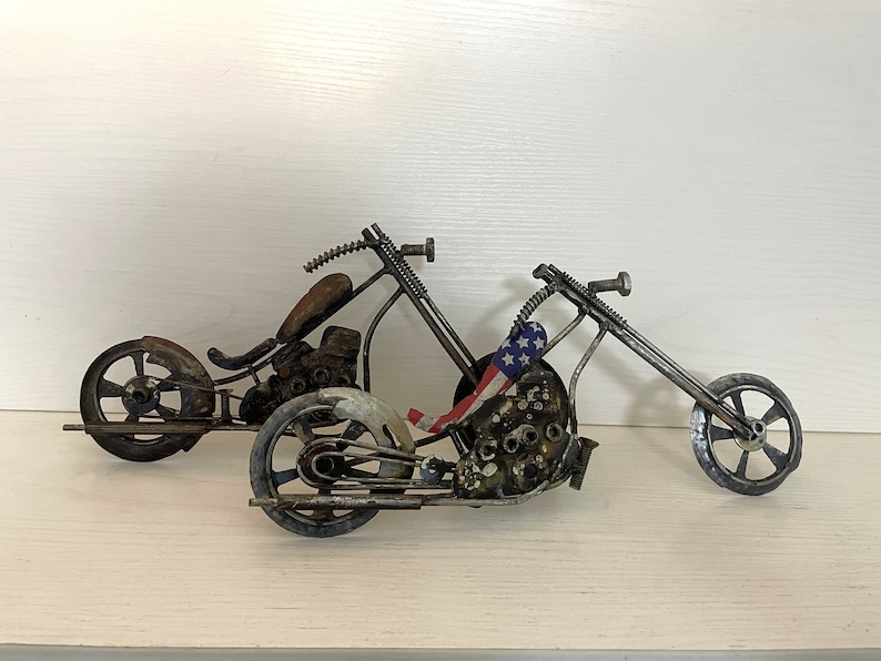 Op de afbeelding: Twee handgemaakte motorfiets-sculpturen van metaal, een met een Amerikaans vlagontwerp. De motorfietsen zijn gemaakt van gerecyclede metalen onderdelen en hebben een rustieke, industri&euml;le uitstraling.