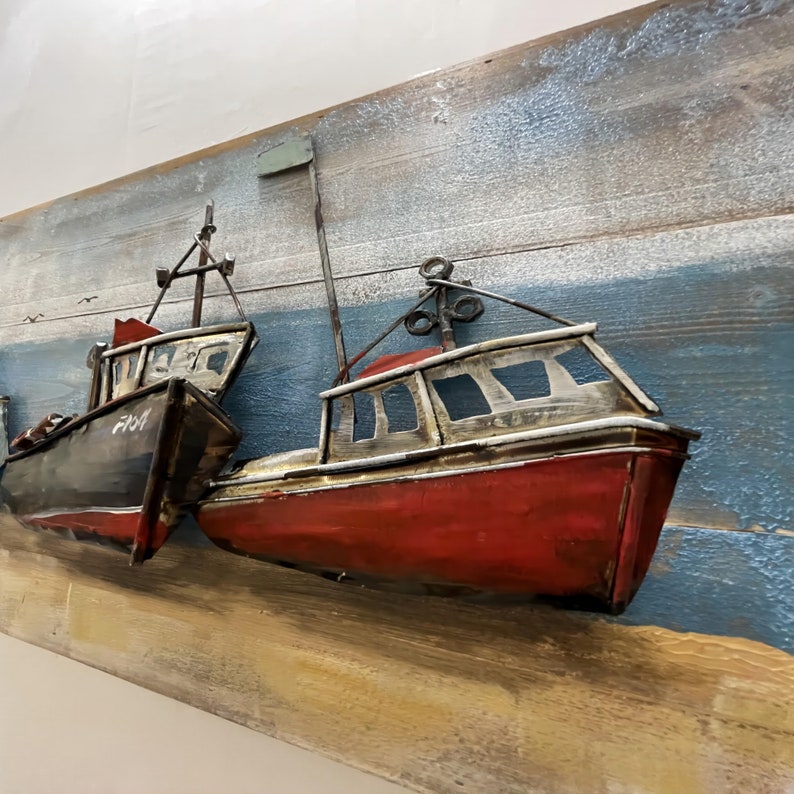 Peut inclure: Une sculpture murale en m&eacute;tal repr&eacute;sentant trois bateaux de p&ecirc;che rouges et noirs sur un fond en bois avec un ciel peint en bleu et blanc. Les bateaux sont superpos&eacute;s et semblent &ecirc;tre en trois dimensions.