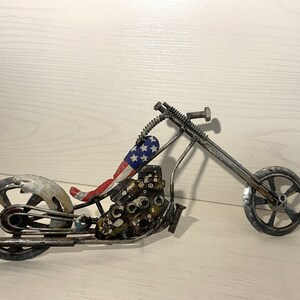 Op de afbeelding: Een metalen motorfiets sculptuur met een rood, wit en blauw Amerikaans vlag ontwerp op de stoel. De motorfiets is gemaakt van gerecycled metaal en heeft een rustieke, industri&euml;le uitstraling.