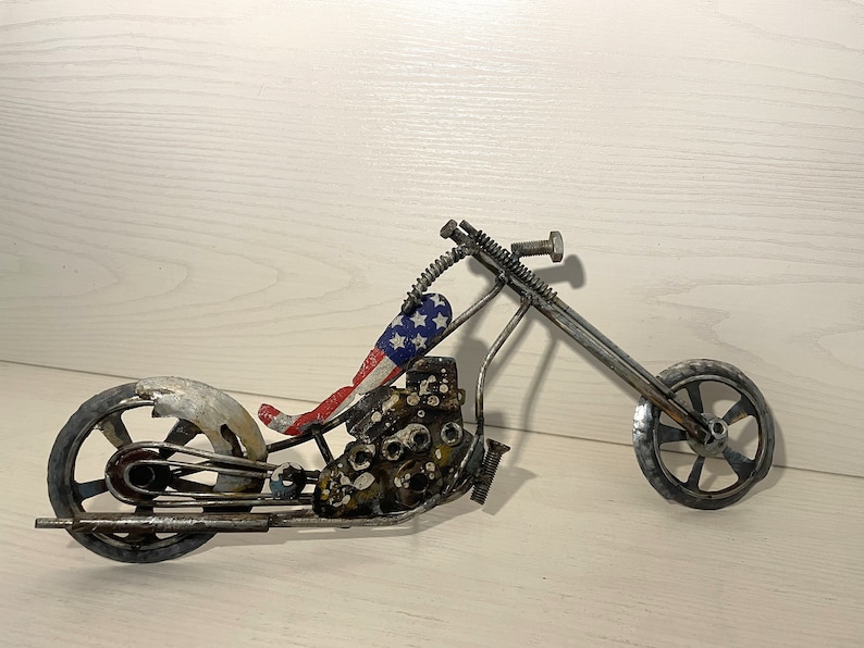 Op de afbeelding: Een metalen motorfiets sculptuur met een rood, wit en blauw Amerikaans vlag ontwerp op de stoel. De motorfiets is gemaakt van gerecycled metaal en heeft een rustieke, industri&euml;le uitstraling.