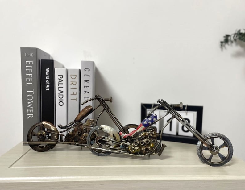 Op de afbeelding: Een metalen sculptuur van een motorfiets met een rood-wit-blauw ontwerp op de stoel. De motorfiets is gemaakt van verschillende metalen onderdelen en staat op een houten oppervlak. De sculptuur staat voor een boekenkast met boeken met de titels "The Eiffel Tower", "World of Art", "Palladio", "Drift" en "Cereal".