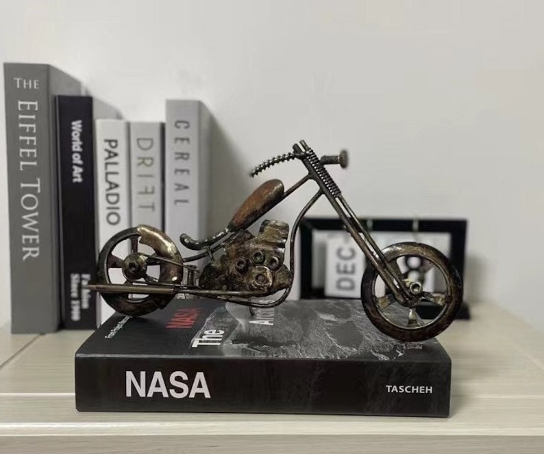 May include: A metal motorcycle sculpture with a brown, rusted finish. The motorcycle is made of various metal pieces, including nuts, bolts, and screws. The motorcycle is sitting on a black book with the word "NASA" printed on the cover.