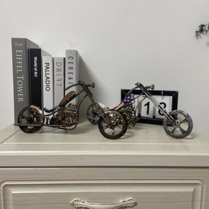 May include: Two metal motorcycle sculptures, one with an American flag design, on a white surface with books in the background.