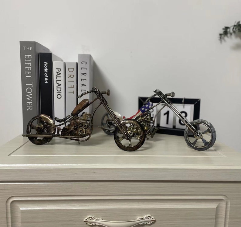 May include: Two metal motorcycle sculptures, one with an American flag design, on a white surface with books in the background.