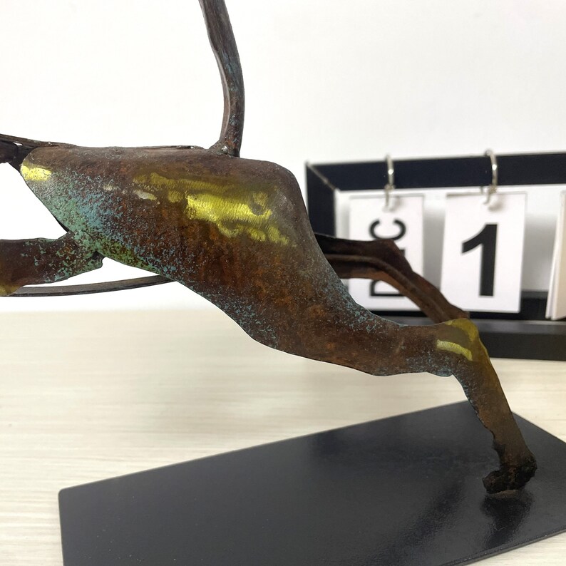 Op de afbeelding: Een roestig metalen sculptuur van een springend dier met een gouden en groene patina. Het sculptuur staat op een zwarte basis en bevindt zich voor een kalender met de datum "21" zichtbaar.