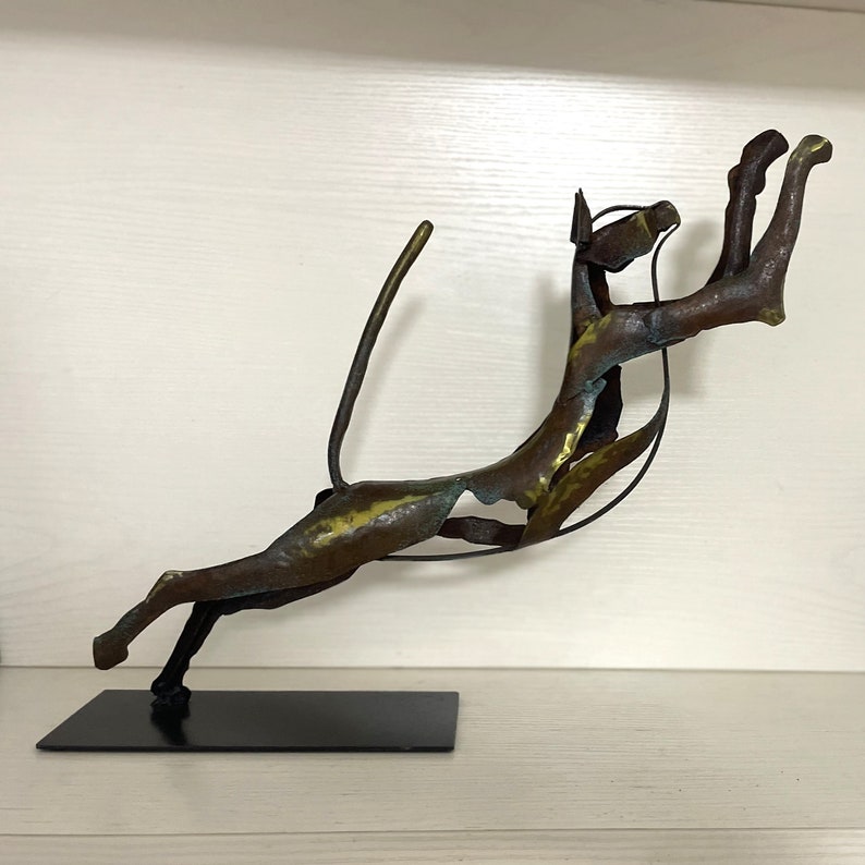 Op de afbeelding: Een bronzen metalen sculptuur van een springende kat met een lange staart. Het beeld is gemonteerd op een zwarte metalen voet.