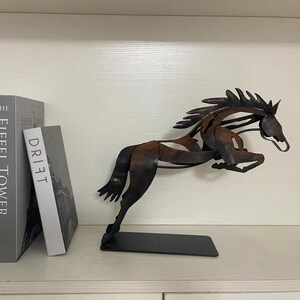 May include: A metal sculpture of a horse in a leaping pose. The horse is made of dark brown and black metal and has a rustic finish. The sculpture is on a black base and is displayed on a shelf with two books.