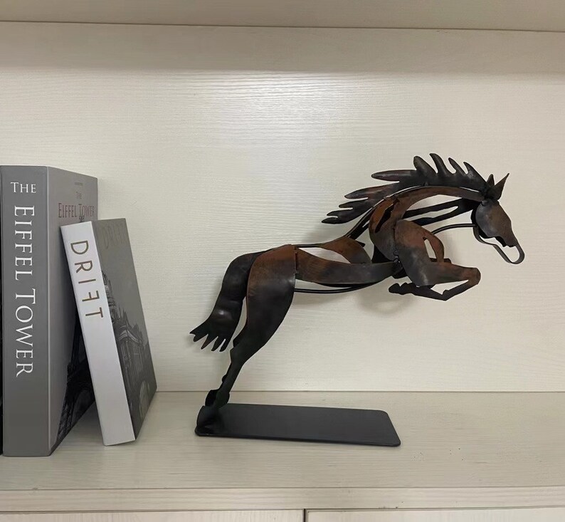 May include: A metal sculpture of a horse in a leaping pose. The horse is made of dark brown and black metal and has a rustic finish. The sculpture is on a black base and is displayed on a shelf with two books.
