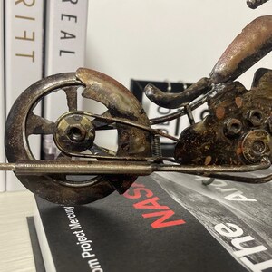 May include: A rusty metal motorcycle sculpture with a detailed design. The motorcycle has a single front wheel and a detailed engine. The sculpture is sitting on a book with the word "NAS" visible.