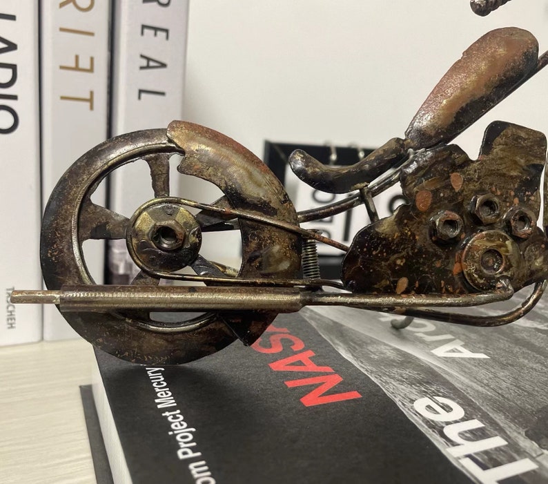 May include: A rusty metal motorcycle sculpture with a detailed design. The motorcycle has a single front wheel and a detailed engine. The sculpture is sitting on a book with the word "NAS" visible.
