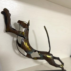 Op de afbeelding: Een metalen sculptuur van een springende kat, met een gouden en groene patina. De kat is geposeerd met zijn achterpoten gestrekt en zijn voorpoten omhoog. Het beeld is gemonteerd op een zwarte basis.