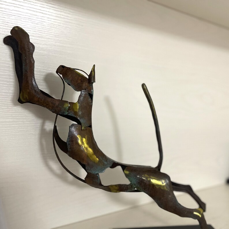 Op de afbeelding: Een metalen sculptuur van een springende kat, met een gouden en groene patina. De kat is geposeerd met zijn achterpoten gestrekt en zijn voorpoten omhoog. Het beeld is gemonteerd op een zwarte basis.