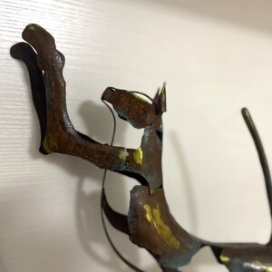 Op de afbeelding: Een roestig metalen sculptuur van een kat met gouden en blauwe accenten. De kat staat in een speelse houding met zijn staart opgerold.