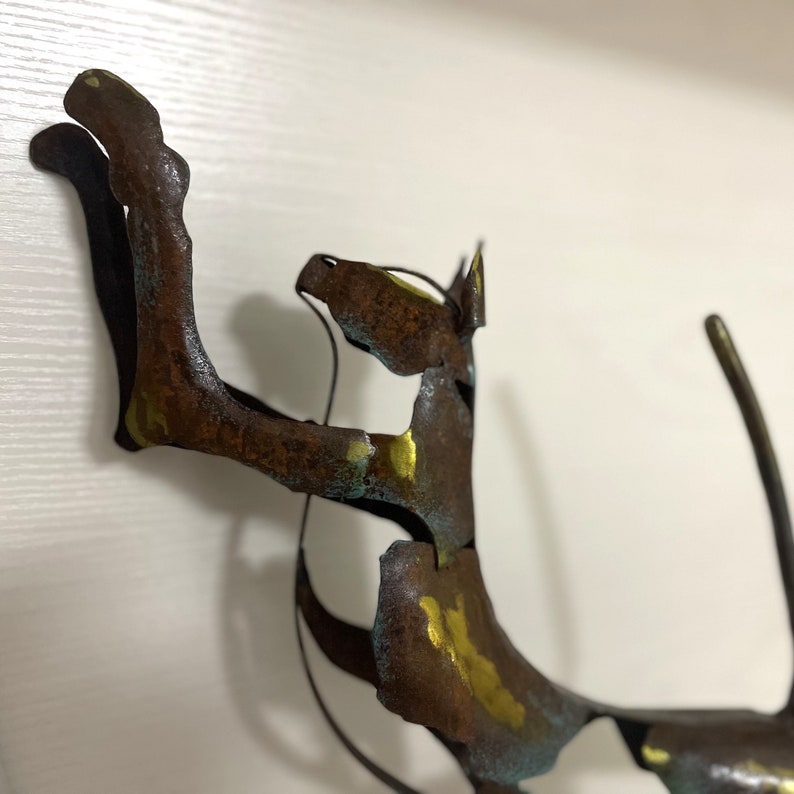Op de afbeelding: Een roestig metalen sculptuur van een kat met gouden en blauwe accenten. De kat staat in een speelse houding met zijn staart opgerold.