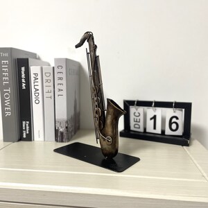 May include: A metal saxophone sculpture in a rustic brown finish with a black stand. The sculpture is on a wooden surface with a black calendar that reads "DEC 16".