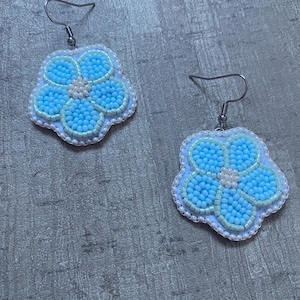 Puede incluir: Un par de pendientes hechos a mano con un diseño floral. Cada pendiente tiene una flor de cuentas azul claro con un centro y borde de cuentas blancas, suspendida de un gancho plateado.