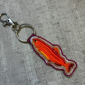 May include: A fish-shaped keyring featuring intricate beadwork in shades of orange, yellow, and red. The fish design is outlined with white and gold beads, attached to a silver-toned metal split ring and a lobster claw clasp for easy attachment.