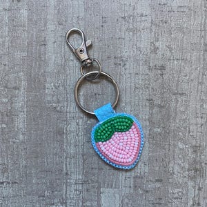 May include: A strawberry-shaped keyring with a silver clasp. The design is made of beads, with pink beads forming the fruit, green beads for the leaves, and a light blue border.