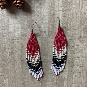 May include: A pair of beaded earrings with a red, black, and white pattern. The earrings are teardrop-shaped and feature a fringe design. The beads are strung together to create a geometric pattern. The earrings have silver-colored hooks.