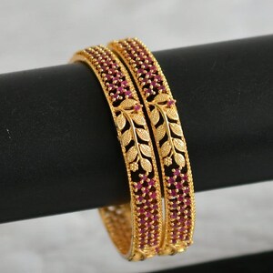 May include: A pair of gold bangles with intricate leaf designs and pink gemstones. The bangles are stacked on top of each other.