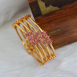 May include: Gold-toned bangles with floral accents. Each bangle features a series of small, vibrant red and green gemstones arranged in a floral pattern. The bangles are stacked together, showcasing their design and craftsmanship.