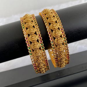 May include: Two gold-toned bangles featuring a central row of circular embossed medallions. Each bangle is bordered by alternating small red, green, and clear stones set in a metallic frame, creating a detailed and textured appearance.