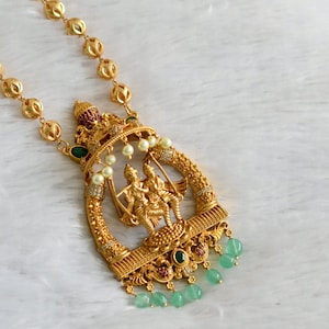 Könnte beinhalten: Eine goldene Halskette mit einem Anhänger, der ein Paar auf einer Schaukel zeigt. Der Anhänger ist mit grünen und weißen Steinen verziert. Die Halskette besteht aus goldenen Perlen.