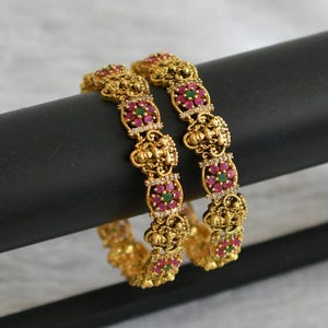 May include: A pair of gold bangles with intricate designs, featuring pink and green gemstones. The bangles are adorned with floral motifs and small, detailed figures.