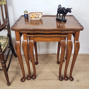 May include: A set of three nested wooden tables with curved edges and cabriole legs featuring carved shell details. On the top table sits a small blue and white cylindrical container, a rectangular floral patterned bowl, and a black cow figurine.