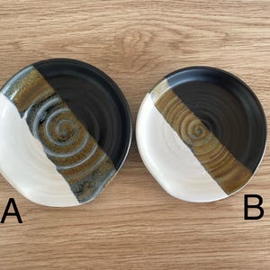 May include: Two handmade ceramic plates with a unique design. Each plate features a diagonal split with a white, black, and brown color scheme. The brown section has a spiral pattern. The plates are labeled A and B.