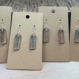May include: Four pairs of handmade earrings displayed on brown cards. Each earring features a rectangular silver frame with a vertical line of small, colorful beads. The bead colors include shades of pink, blue, and brown.