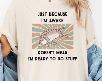 May include: Cream-colored t-shirt with the text "JUST BECAUSE I'M AWAKE DOESN'T MEAN I'M READY TO DO STUFF." The shirt features a graphic of a cat in a relaxed pose, set against a retro-style background with stripes in shades of green, red, and beige.