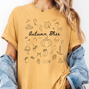 May include: Mustard yellow t-shirt featuring the text "Autumn Bliss" and black line drawings of autumn symbols like pumpkins and leaves. The shirt has short sleeves and a casual fit. The design is printed on the front.