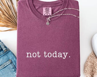 May include: A maroon Comfort Colors t-shirt with the phrase "not today" printed in white. A silver necklace with a sunburst pendant is draped across the shirt. The shirt is folded on a white surface.