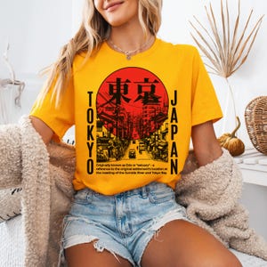 May include: Mustard yellow t-shirt featuring a red and black graphic of a Japanese street scene within a red circle. The words "TOKYO" and "JAPAN" are printed vertically on either side of the graphic. The text at the bottom reads "Originally known as Edo or 'estuary' - a reference to the original settlement's location at the meeting of the Sumida River and Tokyo Bay."