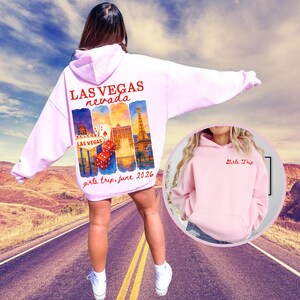 Könnte beinhalten: Ein hellrosa Kapuzenpullover mit dem Text "LAS VEGAS Nevada" in Rot. Auf der Rückseite befindet sich eine Collage von Wahrzeichen von Las Vegas und der Text "girls trip, june 2026". Ein kleineres Bild zeigt einen rosa Kapuzenpullover mit "Girls Trip" in Rot.