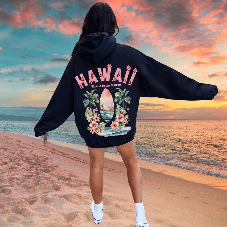 May include: A navy blue hoodie featuring a tropical design with the word "HAWAII" in pink, along with palm trees, flowers, and a beach scene. The text "The Aloha State" is also included.