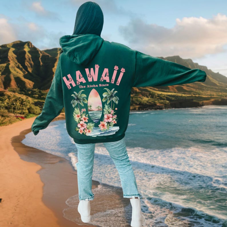 May include: Dark green hoodie with the word "HAWAII" in pink, along with "The Aloha State" text. The back graphic features a surfboard, palm trees, and flowers. The person is wearing light blue jeans and white boots.