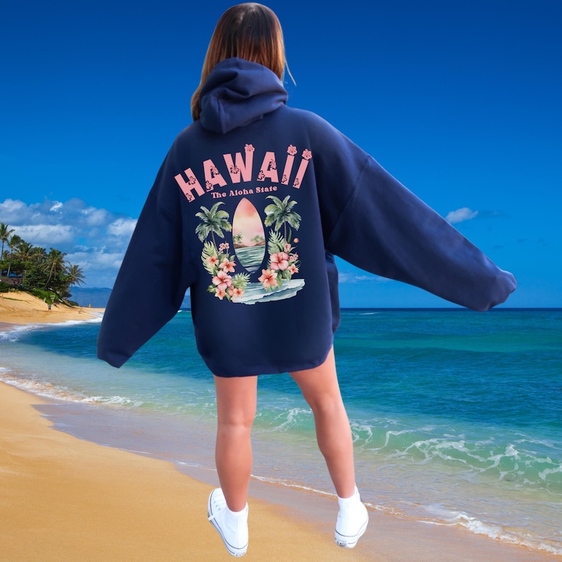 May include: Navy blue hoodie with a graphic on the back that says "Hawaii" in pink letters. The graphic includes a surfboard, palm trees, and flowers. The hoodie is oversized and has a hood.